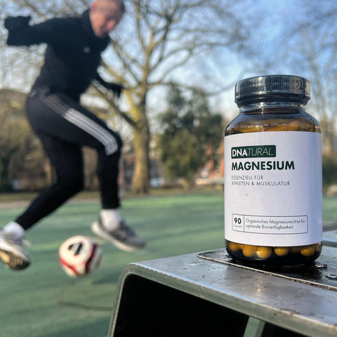 Braunglasflasche DNatural Magnesium auf einem Sportplatz, im Hintergrund ein spielender Sportler – Nahrungsergänzung für aktive Menschen.
